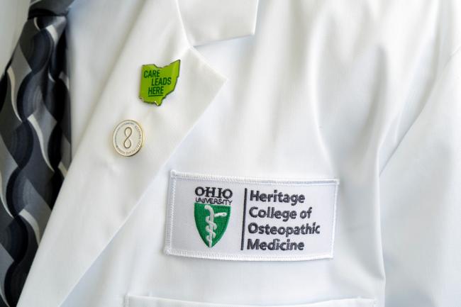 The Heritage College of Osteopathic Medicine logo is shown on a white coat, worn by a doctor. The coat also has a pin that says Care Leads Here