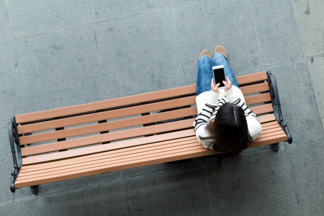 overhead view of a woman alone on a park bench, looking at a cell phone