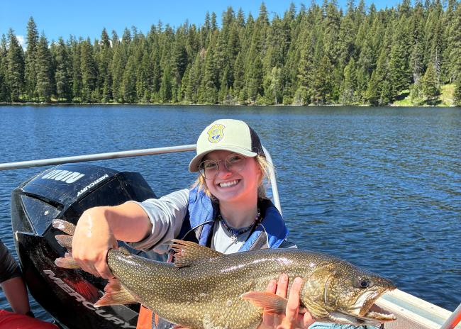 A woman wearing a baseball hat and standing in a boat with pine trees in the background holds a freshly-caught fish out for the camera