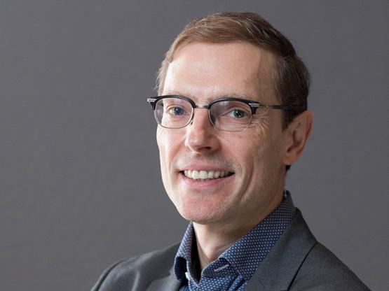  Photo of man wearing glasses, facing viewer, against a neutral grey backdrop. 