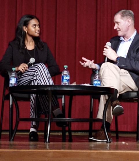  Two people sit side-by-side on a stage, both holding a microphone. The man on the right is speaking directly to the woman on the left 