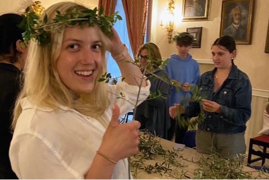  A student wearing a laurel wreath on her head smiles at the camera while others create wreaths in the background 