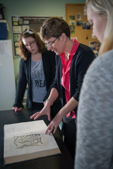  Two students stand with a professor examining a large book open in front of them 