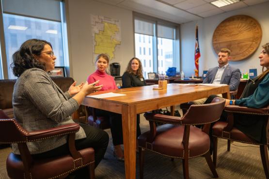 A group of people sit around a conference table, one gesturing as she talks