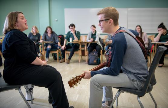  In a classroom, a student holds a guitar while seated across from the instructor as the class observes. 