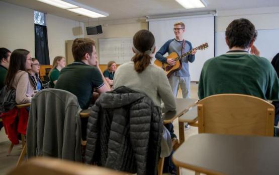  A man stands in front of a classroom full of students and plays the guitar 