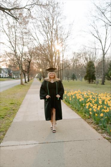  Graduate in cap and gown walking next to daffodils 