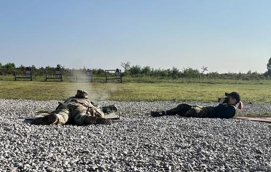  On a gravel surface, a soldier lying on the ground shoots at a target in the near distance while a photographer lies beside him, taking a picture 
