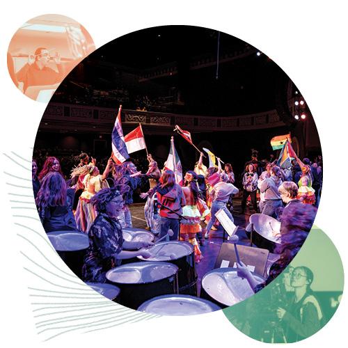 a drummer plays enthusiastically in the foreground while many dancers joyfully wave flags, all on the stage of Templeton-Blackburn Memorial Auditorium; the audience is visible in the far background