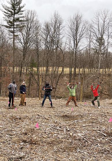 five people pose with shovels in an empty stretch of land, with bare-branch trees in the background