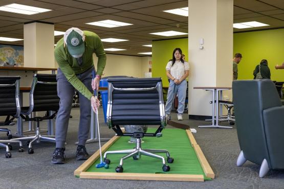 A man plays putt-putt golf, putting around a chair, while an OHIO student looks on in Alden Library