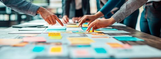  Individuals point at posted notes that are spread out on a table 