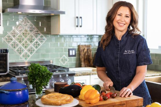  chef christina xenos, wearing a blue work shirt and standing next to a cutting board with vegetables 