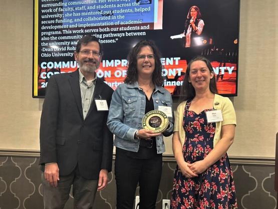  woman holding an award in center of photo featuring 3 people 