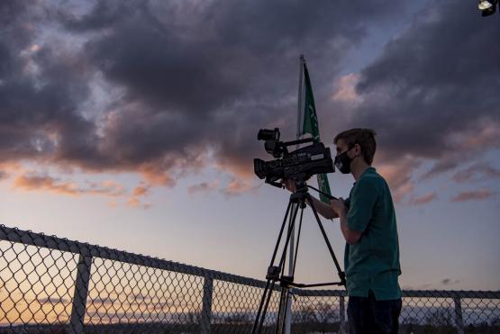  Bryan Kurp stands behind a camera wearing a mask, with a dramatic sunset as background 