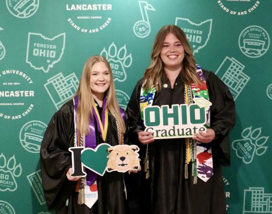  Two OHIO Lancaster graduates are shown in their graduation robes while holding OHIO Graduate signs 
