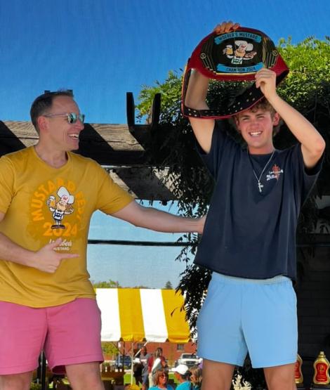 Joey Chestnut points at Jack Legath while they two stand on a stage and Legath holds up a championship belt