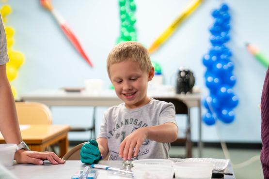  A young student smiles and wears a glove while working on a STEM project at a table, and teachers stand nearby 