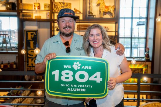 Two OHIO alumni hold a sign that says We are 1804 Ohio University Alumni