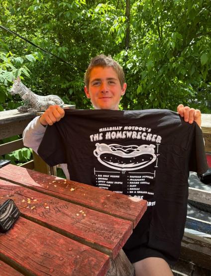 Jack Legath holds up a shirt that says Hillbilly Hotdog's The Homewrecker, while he sits at a table outside