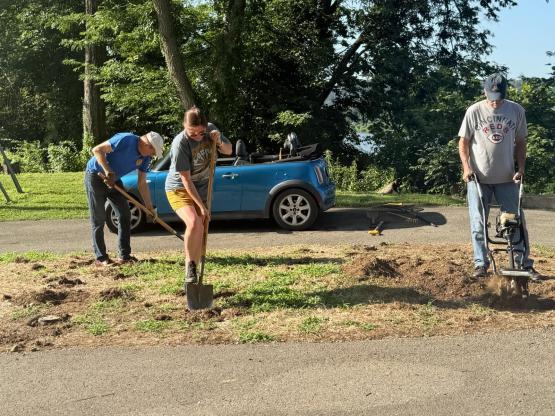  Three people work on outdoor garden space with shovels, rakes, tiller. 