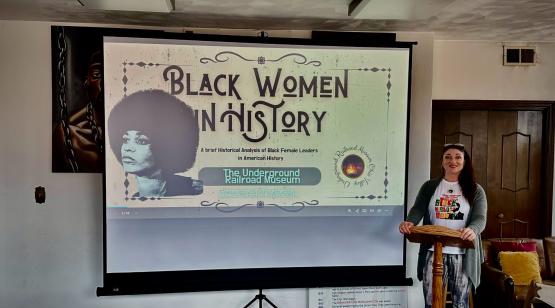  Photo of a female with long black hair standing behind a wooden podium, next to a projection screen that show a slide stating &amp;quot;Black Women in History&amp;quot; 