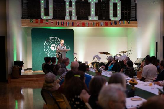President Lori Stewart Gonzalez speaks before a large crowd, and underneath international flags and the word OHIO at the SISAL Dinner