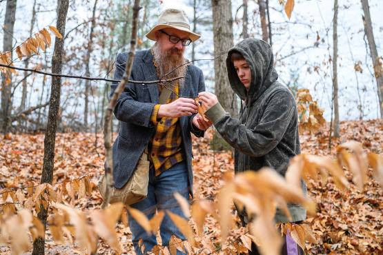  a bearded professor wearing a hat and glasses shows a leaf specimen to a young student wearing a hoodie 