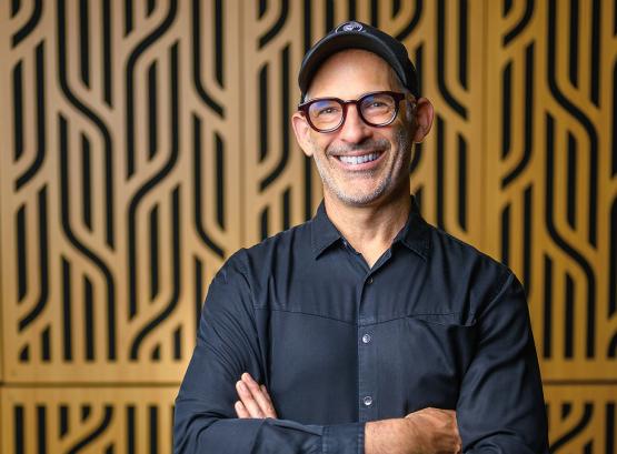  a man wearing a button-down shirt, baseball cap and glasses stands in front of a textured wall, smiling 