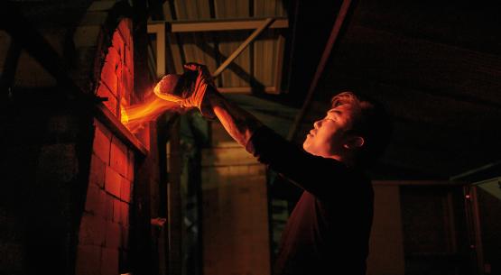  flames shoot from a small gap as a graduate student wearing gloves removes a brick from a kiln to check on its contents 