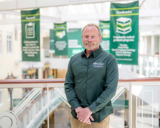 a man with short gray hair and beard in a green Ohio University zip-up shirt poses in front of green banners in a brightly lit atrium