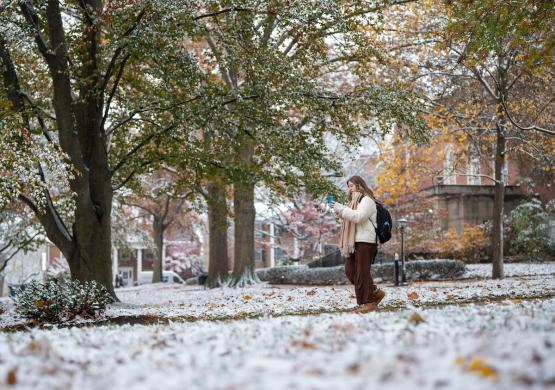 An OHIO student looks at her phone while walking on the College Green, which has light snow on the ground and colorful fall leaves on the trees
