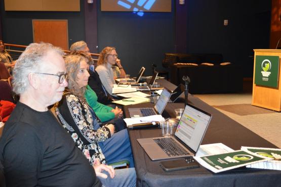 The judges sit at a table with laptop computers in front of them and watch the presentations at the Eco Challenge