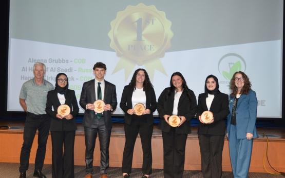 The winners of the Eco Challenge, along with advisors, hold trophies while standing on stage at the Eco Challenge, and a 1st Place graphic is shown behind them