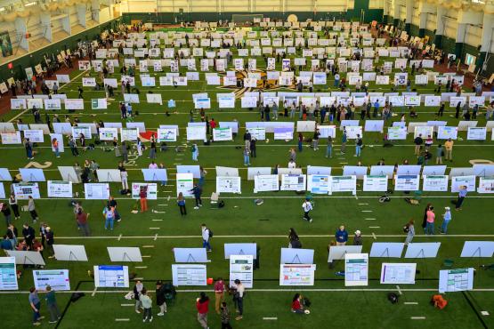 Rows of research posters, with students standing near them, are shown in the Walter Fieldhouse for the Student Research Expo.