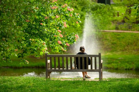 An OHIO student relaxes on a park bench while water from the pond sprays into the air at Emeriti Park on a spring day