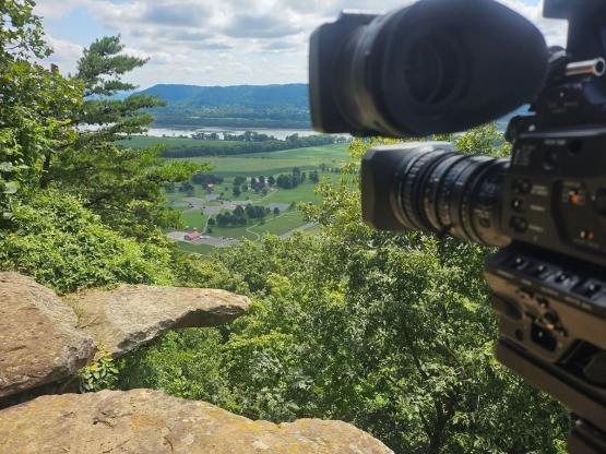 A video camera is shown from behind as it overlooks a green valley and river below, while green trees are on the hillside to the left