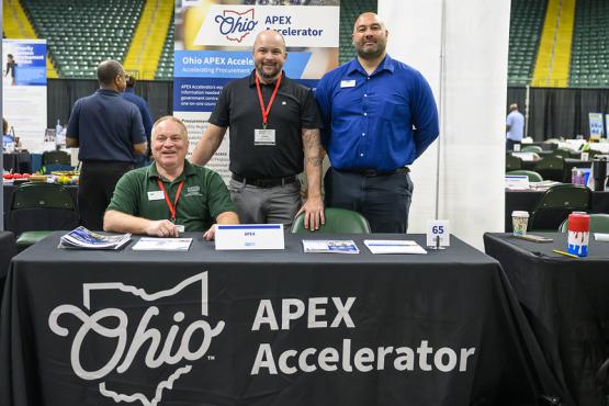 Three individuals sit and stand behind a table that has an Ohio APEX Accelerator banner on it at the Ohio Business Matchmaker event