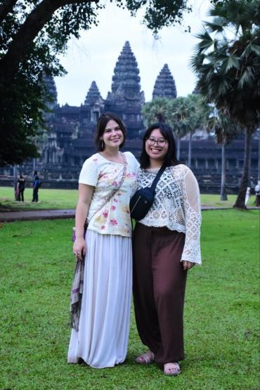 Andrea Tiu and Callia Fout stand together outside in front of green grass and old buildings in Cambodia