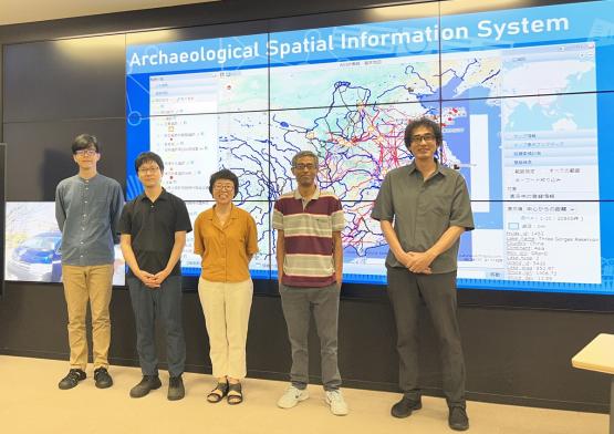 OHIO and Chubu University representatives stand in front of a video screen that has the words "Archaeological Spatial  Information  System" across the top