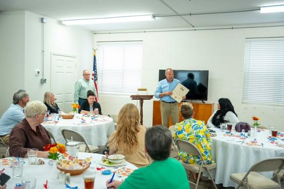 A speaker gives a presentation while  members of the Mayors' Partnership for Progress sit nearby at tables and listen
