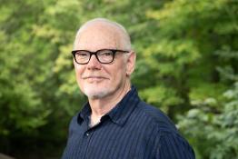 With a background of green leaves, a man wearing black-rimmed glasses faces the camera