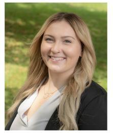 Headshot of Maya Clouse Henry smiling and wearing a gray shirt and dark suit jacket with an grassy outdoor background 