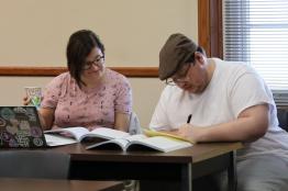 Students studying at a desk, male is writing, female is observing