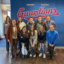 a group of 10 students stand in front of a wall with the Cleveland Guardians logo