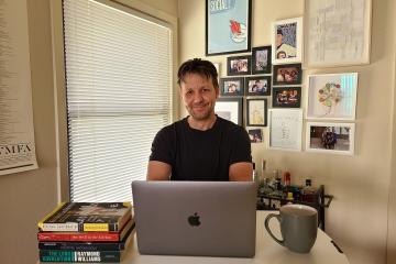Denis Flaschner sits behind a desk with a stack of books, Apple laptop, mug, with a window and wall of pictures and artwork behind him