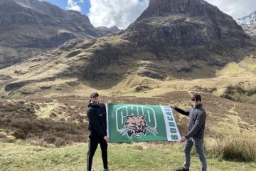Two people hold up an Ohio University athletics flag in front of a hilly landscape