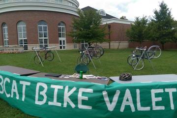 A table set up with a green cloth hanging over it that says "BOBCAT BIKE  VALET" with several bikes on the lawn behind it, with Walter Hall in the background