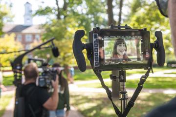 A camera operator is holding a camera screen on College Green, with the screen focused on a smiling student