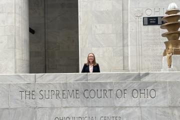 Kirsten Thomas poses behind a marble wall that reads "Supreme Court of Ohio" with below it "Ohio Judicial Center"